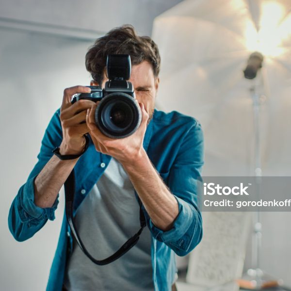 In the Photo Studio with Professional Equipment: Portrait of the Famous Photographer Holding State of the Art Camera Taking Pictures with Softboxes Flashing in Background.